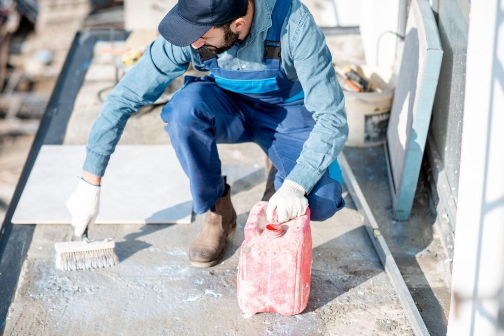 Workman in uniform priming concrete with brush for tiles lying on the balcony