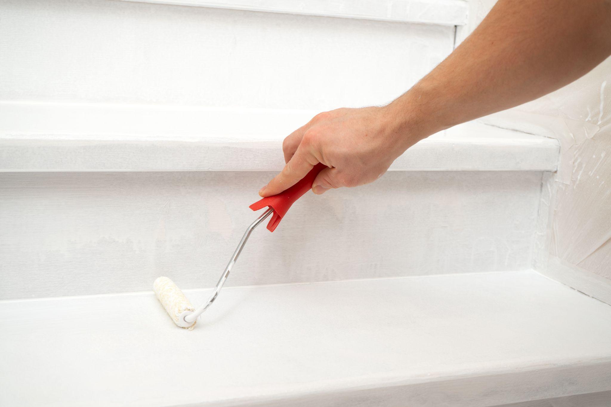 Hand paints a wooden staircase white with paint and a roller.